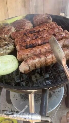 Here’s a description of the video:
The video shows food being cooked on a grill. There are several long, thin sausages, patties, a slice of onion, and some green peppers on the grill, which is surrounded by smoke. The sausages are turning brown and appear almost cooked. Metal tongs are used to adjust the sausages on the grill. The grill itself is black and sits on a metal stand. The background consists of a wooden wall. The camera angle is close-up, focusing on the food and the grilling process.