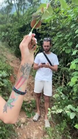 The video captures an outdoor scene in a lush, green, hilly environment, consistent with Nậm Dịch Commune, Vietnam. In the foreground, a person's tattooed arm and hand with light-colored painted fingernails holds up a leafy branch. A bearded man, wearing a white t-shirt, light shorts, and sandals, with a black cross-body bag, stands on a dirt path in the mid-ground. He is initially seen looking at a smartphone, then looks up, smiles broadly, laughs, makes a playful face, and waves at the camera. In the distant background, another person, possibly a woman wearing a red top, is visible standing on a grassy hillside. The setting is characterized by abundant green foliage and trees, indicating a natural, possibly rural or mountainous, area. The man's expressions convey humor and playfulness, suggesting a lighthearted moment during an outdoor excursion or hike. The lighting suggests it is daytime.