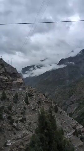 The video shows a mountainous landscape with a cloudy sky. In the foreground, there is a steep, rocky slope covered with sparse vegetation. A road is visible carved into the side of the mountain, with a few vehicles on it and a small building next to the road. In the background, more mountains rise, some peaks obscured by clouds. Power lines are strung across the frame. The overall impression is a remote, rugged environment.