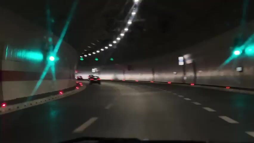 The video shows a point-of-view shot from inside a car driving through a tunnel. The tunnel has white walls and a dark ceiling with numerous lights. There are green directional arrows illuminated on the ceiling, indicating the direction of travel. The road is paved and marked with white dashed lines. Other cars are visible ahead, driving in the same direction. Red lights are positioned along the sides of the road. The lighting in the tunnel creates reflections on the car's windshield.