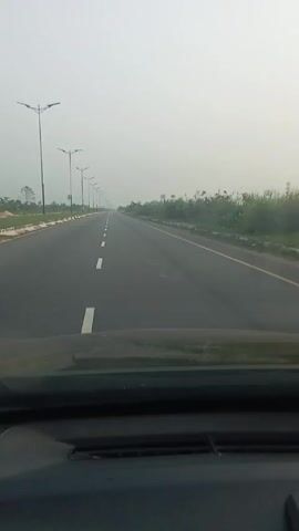 The video shows a view from inside a vehicle driving on a straight, multi-lane highway. The highway is lined with street lights. On either side of the road, there is vegetation, including bushes and some banana trees. The sky is overcast, creating a foggy or hazy atmosphere. The dashboard of the vehicle is visible at the bottom of the frame.