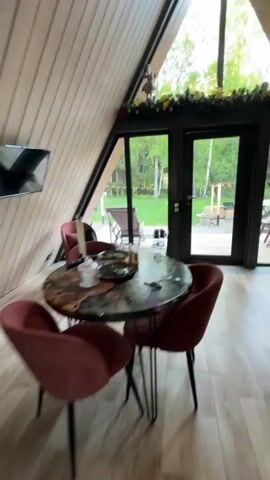 The video starts indoors, showing a living area with a triangular window above a glass door. The interior features a round table with red chairs and a light-colored wood floor. The camera moves towards the glass door, then a hand opens it, revealing an outdoor patio made of wood. The patio overlooks a grassy area with trees.  A hot tub is visible, surrounded by decorative arrangements, and there is also a grill area nearby. The view is peaceful and scenic, with the trees suggesting a woodland setting.