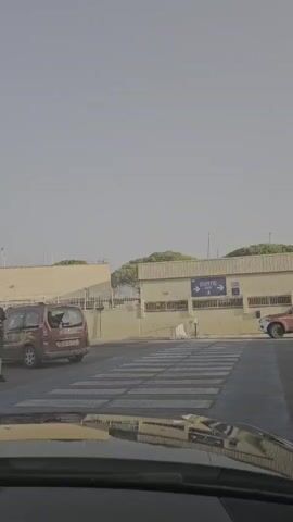 A security guard in a black shirt and shorts, wearing a cap, directs traffic at a crosswalk. A red van is stopped at the crosswalk, and another car is approaching from the left. In the background, there is a building with the word "SORTIE" and an arrow pointing to the right. The sky is clear and blue, and the sun is shining.
