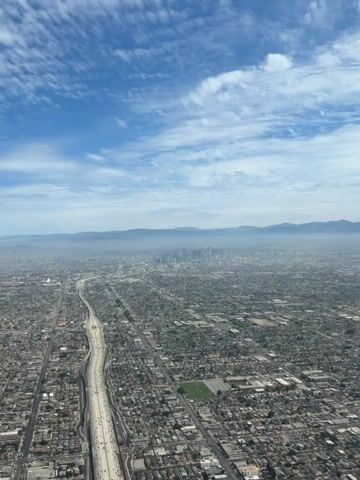 An aerial view shows a sprawling city with a river and highway cutting through it. In the distance, a cluster of skyscrapers represents the downtown area, partially obscured by haze. Rolling hills or mountains form the backdrop, with a clear blue sky dotted with wispy clouds above. The urban landscape extends as far as the eye can see, characterized by a dense network of residential buildings and commercial structures.
