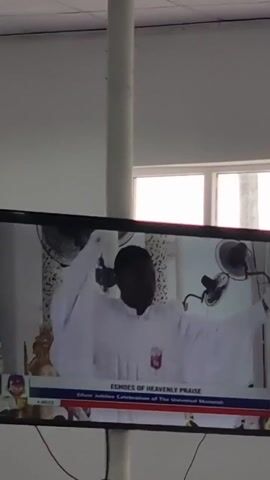 A television screen shows a man wearing white robes speaking into a microphone.  The screen displays a graphic that reads "ECHOES OF HEAVENLY PRAISE Silver Jubilee Celebration of The Universal Monarch." A shot of a large crowd, most of whom are also wearing white robes, is also seen on the screen. The television is mounted on a wall in a white room with a tall white pillar and a window. Cables are seen running down the wall from the television.