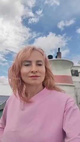 A woman with pink hair and a pink sweatshirt is on the deck of a boat. She is looking at the camera. The sky is cloudy.