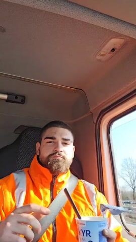 The video shows a man eating yogurt in the cab of a truck. The man has dark hair and a beard. He is wearing an orange high-visibility safety jacket and a seatbelt. He holds a plastic container of KYR skyr yogurt in one hand and a spoon in the other. He eats a spoonful of yogurt and then looks out the window. The truck is moving on a highway. The sky outside is clear and blue. The interior of the cab is simple with a light grey headliner and visible controls.