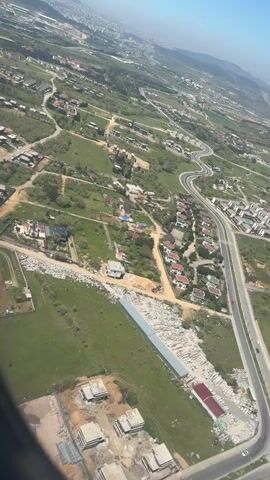 The video shows an aerial view of a suburban area. The landscape is a mix of green fields and residential areas with houses, streets, and roads. A highway with multiple lanes winds its way through the terrain. The camera captures the scene from above, likely from an airplane window. The sky is clear with a slight haze in the distance. The overall scene appears serene and peaceful.