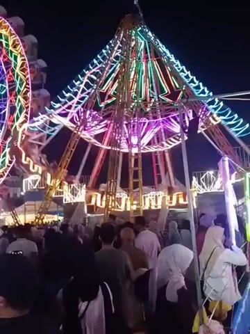 The video shows a crowded amusement park at night. In the background, a large Ferris wheel illuminated with colorful lights is visible, along with another carnival ride with similar lighting. A dense crowd of people is gathered in the foreground, with many looking towards the rides. Some individuals are holding children. The atmosphere appears festive.