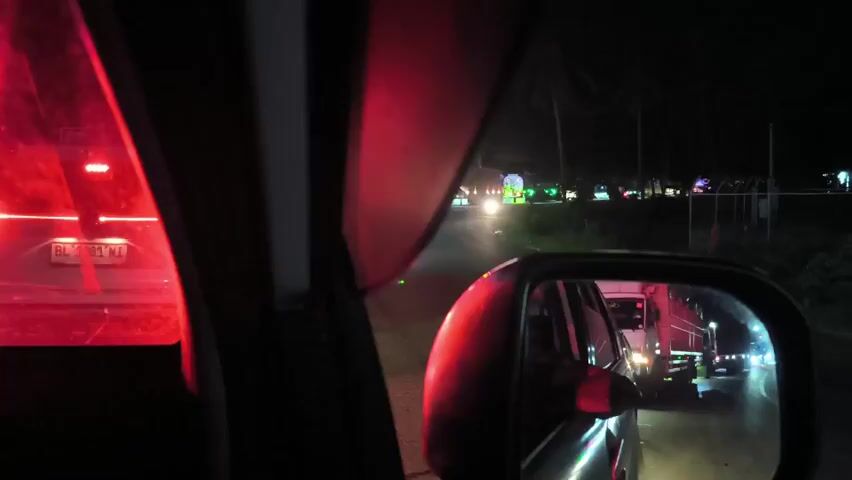 The video shows the perspective from inside a car at night, likely in slow-moving or stopped traffic. The taillights of the car ahead are brightly illuminated. In the side mirror, a truck is visible, its lights reflecting. A row of lights, possibly from a festival or event, can be seen in the distance along the road. The overall environment is dark, with the primary illumination coming from the vehicle lights.