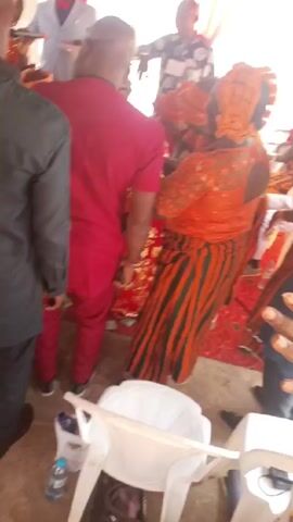 The video captures a vibrant cultural event, possibly a celebration or gathering, based on the traditional clothing and energetic dancing. The foreground features a group of people moving to the music, their bodies only partially visible.  One woman in the center is dressed in an orange lace top, an orange and dark green striped skirt, and a matching orange headwrap with a maroon accent. To her left, a man in a red suit and black shoes stands observing the scene. The camera angle occasionally shifts, showing more of the crowd and revealing more details such as the presence of a white plastic chair and a water bottle on the floor. Another man in a long grey tunic is seen interacting with the man in red. The atmosphere is lively, with people interacting, laughing, and moving to the beat.