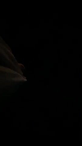 The video begins with a close-up shot of a person's hand in dark, possibly outdoor, terrain. The hand is reaching down towards something on the ground. The terrain appears to be rocky or uneven. The shot then shifts to a wider view, showing the person's legs and feet as they walk or stand in the dark. There is a faint light source visible in the distance, possibly a moon or street light, illuminating a patch of dark blue sky and a rugged surface that could be rocks or a ledge. The scene is mostly dark and indistinct.