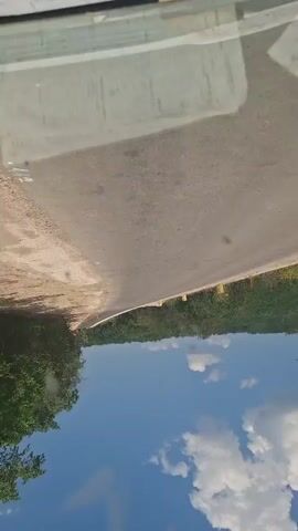 The video shows an upside-down view of a road construction site. The foreground is dominated by the road surface, which appears to be unfinished with gravel and dirt. On the left side of the road, there are trees and lush green foliage. On the right side, the foliage continues, with yellow poles marking the edge of the road. In the distance, there are visible signs of construction, including orange traffic cones and what appears to be construction equipment. The sky is blue with scattered white clouds, suggesting it is daytime and the weather is clear. A reflection on what seems to be the car's windshield shows some text, but it's not legible. The overall scene suggests an ongoing road construction project in a rural or semi-rural area, possibly in Hîncești, Moldova, given the location context. The upside-down perspective creates a disorienting effect.