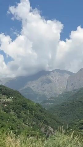 The video shows a scenic mountain view with lush green hills and majestic peaks partially covered by clouds against a partly sunny, blue sky.