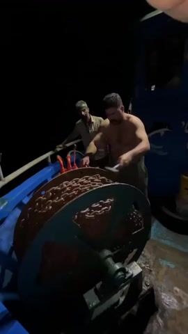 Two shirtless men are on the deck of a boat at night. They are operating a large winch with a heavy chain wound around it. The man in the foreground is operating the winch with a lever, while the man in the background stands behind him and appears to be guiding the chain. The boat is dark except for the lights illuminating the winch and the men. There is a railing to the left and the side of the boat on the right is blue with painted dolphins.