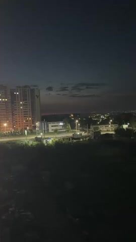 The video begins by showing a wide shot of a cityscape at dusk. A tall, illuminated apartment building is visible on the left side of the frame. In the distance, faint lights of the city can be seen. As the video progresses, the camera zooms in on a section of the cityscape. A few scattered clouds are present in the darkening sky. In the middle distance, there are several low-rise buildings, some with illuminated windows and streetlights. Trees and dark, shadowy areas indicating vegetation are also visible. The most prominent feature that develops in the video is a series of colorful fireworks exploding in the sky over the distant city. The fireworks display various colors, including yellow, pink, and green, against the dark background. The video captures the fleeting moments of the fireworks as they illuminate the night sky.