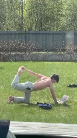 A shirtless man wearing gray sweatpants does yoga on the grass. He kneels on his left knee with his right leg bent upward and grasping his right foot with his right hand. His left hand is on the ground. A laptop computer, a can, and a black jacket are on the grass in front of him. A green lawn and a dark gray fence are in the background. Trees are visible beyond the fence. The scene is illuminated by natural sunlight.