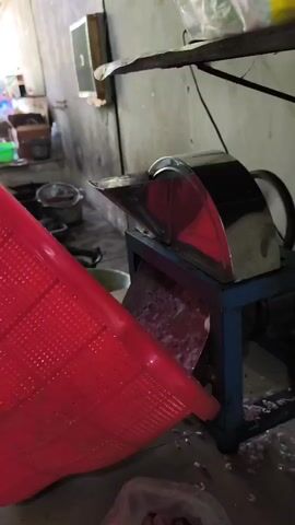 The video shows a red plastic basket being tilted over a stainless steel machine with a blue base, presumably to feed chopped onions into it. The machine is actively processing the onions, which are visible falling out of the machine into what appears to be a white plastic bag on the floor. The background suggests a kitchen or food preparation area with shelves, containers, and electrical outlets.