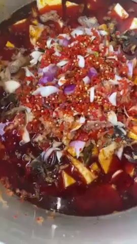 A close-up shot depicts a large, stainless steel pot filled with a dark, reddish-brown liquid. Scattered throughout the pot are cubed pieces of a light yellow food, possibly tofu, along with diced red onions, tomatoes, and a generous amount of chopped red chili peppers and flakes. A hand with light blue nail polish is visible holding a large spoon, stirring the mixture. The contents of the pot appear to be in the process of being mixed together, suggesting the preparation of a spicy, possibly Asian-inspired, dish.