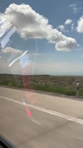The video shows a view from inside a vehicle, looking out the window. The window reflects the interior of the vehicle, including what appears to be a person wearing a white shirt. Outside the window, there is a road, a grassy field, and a blue sky with white clouds. The sun is shining, and there are some red lens flares visible on the road. The video is shaky, suggesting the vehicle is in motion.
