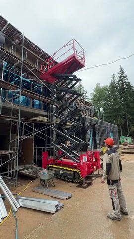A construction worker in a hard hat and work clothes stands near a red and black tracked scissor lift that is extended upwards. The scissor lift is positioned in front of a building under construction. The building has scaffolding around it and appears to be unfinished, with exposed framing and windows. In the foreground, there are piles of building materials, including metal beams and a wheelbarrow. The ground is dirt. Trees are visible in the background. The sky is overcast.