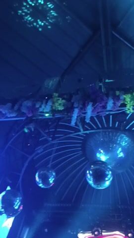 The video shows a nightclub or event venue with blue lighting and disco balls hanging from the ceiling. There are floral decorations and a digital display with text. People are present, some dancing and others standing around. The camera moves, showing different parts of the venue and the crowd.