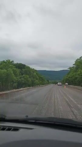 The video shows a view from inside a vehicle driving on a paved road. The road appears to be under construction or repair, with areas of loose gravel and visible tire tracks. Dense green trees line both sides of the road, and rolling hills are visible in the distance under a cloudy sky. Ahead on the road, there are two cars, a white sedan and a darker vehicle, and a person standing in the middle of the road. An orange traffic cone is also visible on the right side of the road. The weather appears overcast.