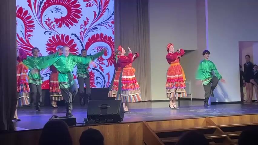 On a wooden stage under the bright lights, a group of young dancers perform a traditional folk dance. The stage backdrop is a screen displaying a bold red and white floral pattern. The dancers, a mix of boys and girls, are dressed in colorful costumes. The boys wear green tops and dark pants, while the girls are in red tops and long patterned skirts. Some dancers wear headdresses. They move in coordinated steps, occasionally raising their arms and changing positions. In the foreground, part of the audience is visible, their figures silhouetted. There is a speaker on the stage. A doorway is visible in the background.