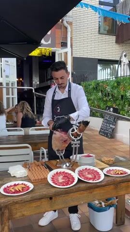 A man in a white shirt, black apron, and black gloves is slicing a large ham on a wooden table outdoors. He is wearing black pants and white sneakers. The ham is secured in a metal stand. In front of the ham are several plates of sliced ham, a wooden block with knives, and other serving dishes. Behind him, people are seated at tables under an umbrella, and a building with greenery is visible. A sign with the word "ESCAPER" is posted on the right.