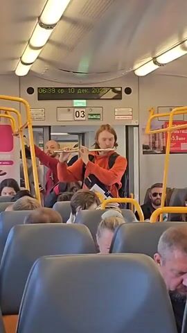 A man with long blonde hair, wearing an orange hooded sweatshirt and a black bag across his chest, plays a silver flute. He is standing in a train car, facing forward. The train car appears to be a commuter train, with rows of seats visible in the foreground. Other passengers are seated and looking in various directions, some appear to be looking at the musician, others are looking away. Behind the musician, a sign displays the time 06:39, the day of the week sr (likely Sreda, Wednesday in Russian), the date 10 дек. (December 10), and the year 2025. Another sign above a door indicates '03' and 'ВЫХОД' (Exit). The lighting in the train car is bright, with fluorescent lights overhead. Yellow handrails are visible, providing support for standing passengers. The overall impression is of a public transportation setting with an impromptu musical performance.