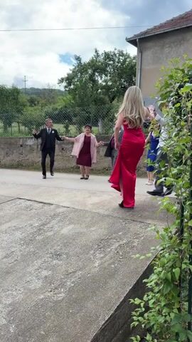The video captures a wedding celebration on what appears to be a concrete road in a rural setting. A bride in a white, long-sleeved gown adorned with lace and a floral headpiece is seen dancing. She is joined by others dancing in a line. A man in a dark suit and a woman in a maroon dress are holding hands and dancing, as well as another man in a gray suit and a woman in a blue dress. Another woman, wearing a long red dress, is also part of the line. The background includes a building, a metal fence, and lush greenery, including a climbing plant that partially obscures the view on the right side. In the distance, there is a hilly landscape under a cloudy sky.