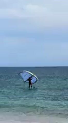 A person is seen in the water, standing on a wing foil board and holding a large, light blue and white wing. The water is a clear blue-green, and the sky above is overcast with light clouds. The horizon line is visible in the distance, separating the sea from the sky. The person is facing slightly left, adjusting the wing as if preparing to catch the wind or just having landed in the water. The sound of wind and waves is audible, suggesting a breezy day. The activity is taking place on the open sea, near the coast of Barbate, Spain. No text is clearly visible on the equipment.