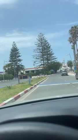 The video shows a first-person view from inside a car driving along a street in Loualidia, Morocco, during the daytime. The weather is sunny with a clear blue sky and some scattered clouds.

The car is moving on a paved road with a painted blue and white striped curb on the right, and a red, white, and blue striped curb bordering a grassy median on the left. Tall, slender pine trees and palm trees line the street, suggesting a coastal or warm climate.

In the distance, a grey SUV is driving ahead, and another vehicle is parked on the right side of the road. White and light-colored buildings are visible behind the trees and on the right side of the street. Streetlights are also visible along the road. The overall scene depicts a typical, pleasant drive through a sunny town. No distinct text is clearly legible on any signs or objects.