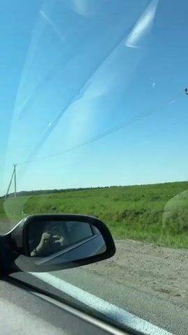 Here is a concise description of the video:

The video shows a view from inside a car driving on a highway. The scenery outside includes green fields, power lines, and streetlights passing by. A person's hand holding a camera is visible in the side mirror. Other vehicles are present on the road. The sky is blue and clear.
