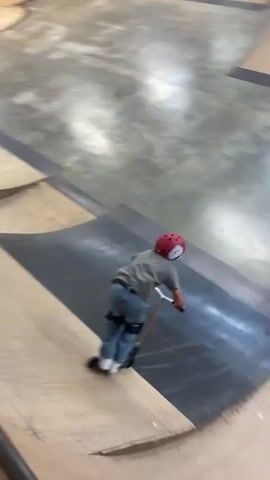 A person on a scooter with a helmet and pads enters a skate park ramp. The person rides up the ramp and performs a trick, appearing to fall. The environment is an indoor skate park with wooden ramps and a high ceiling with lights. Another person is visible in the background filming.