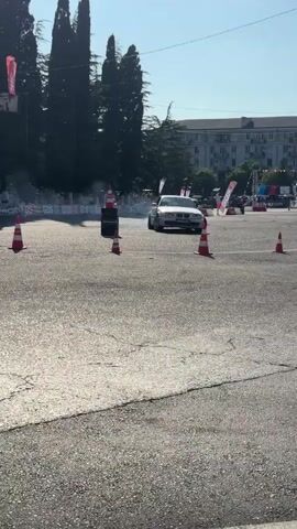 A white sedan is seen drifting around cones on an asphalt surface. The car is kicking up a significant amount of smoke from its tires. The scene appears to be outdoors on a sunny day, with trees and buildings in the background. There are several orange traffic cones placed on the ground, marking a course. The car is the primary focus of the video, demonstrating a skilled maneuver.