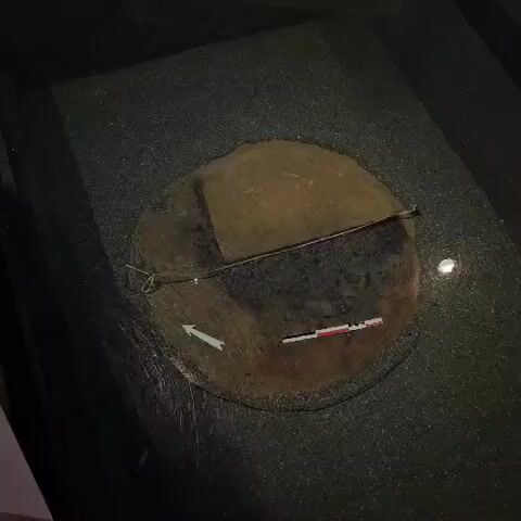 The video shows an aerial view of an archaeological excavation. A circular feature in the ground is visible, with a ruler and a directional arrow placed within it. The feature appears to be some kind of pit or posthole, with darker soil in the center and lighter soil around the edges. There is also a thin wire stretched across the feature. The surrounding ground is dark and granular, possibly dirt or gravel. The lighting is dim, suggesting the excavation is taking place in low-light conditions, possibly underground or at night.
