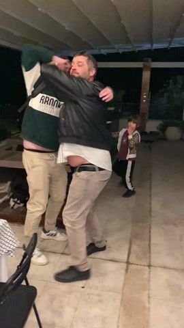 Here’s a description of the video:

The video shows two men dancing together on a patio at night. The men are of similar height. One is wearing a dark green sweatshirt with white lettering, khaki pants, and white sneakers. The other is wearing a black leather jacket, a striped shirt, khaki pants, and black shoes, and has a beard and sunglasses. They are playfully wrapping their arms around each other as they dance. A young girl wearing a bright yellow jacket, black and white track pants, and purple sneakers dances nearby but not with the men. There is a table with a black and white patterned tablecloth, and a black chair. In the background, there is a wooden table and a view of a city lit up at night. The patio is covered by a white roof.