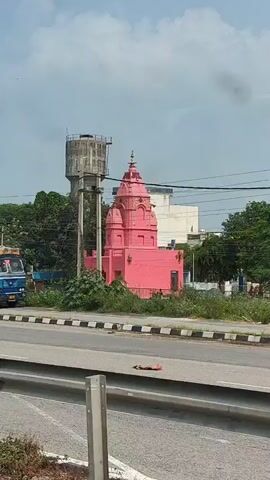 A truck is parked on the side of a road next to a pink Hindu temple. A tall water tower is visible in the background behind the truck. The sky is overcast.