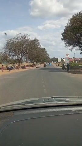The video shows a view from inside a car driving down a two-lane road. There are trees on either side of the road. On the left, people are gathered near the side of the road. Further down the road, there are other vehicles, including a white van and a blue car. On the right side of the road, there is a sign for a gas station. The sky is blue with white clouds.