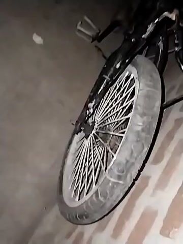 The video shows a black bicycle leaning against a wall. The bicycle has white spokes on the front wheel. There are four coconuts on the ground next to the bicycle. The scene appears to be outside at night. The lighting is dim.
