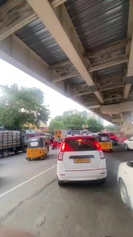 The video shows a congested street scene in what appears to be a city in India, likely during daylight hours. A sky bridge or elevated structure spans overhead, casting shadows on the road below. The road is filled with various vehicles, including cars, motorcycles, auto-rickshaws, and a truck. The vehicles are stopped in traffic, suggesting a traffic jam. Several people are visible on motorcycles, and some are walking on the side of the road. Trees and buildings line the street in the background. The sky is overcast. The overall impression is one of a busy, urban environment with significant traffic congestion.