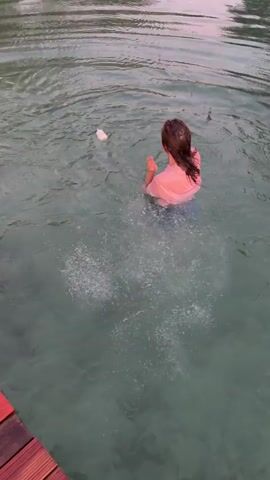 A person with long brown hair, wearing a pink t-shirt and blue shorts, is in the water. The person is splashing the water with their hands and appears to be smiling or laughing. There is a white object floating in the water to the person's left. The water is clear and appears to be a lake or river. Trees or foliage are visible in the background.