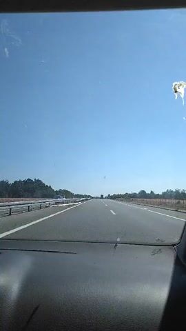 The video shows a first-person perspective of driving on a highway. The road is paved and appears to be a multi-lane highway with a guardrail on the left side. The sky is clear and blue, and trees line the sides of the road. There are some vehicles visible in the distance on the highway. The dashboard of the car is visible in the foreground. There is a small white object in the upper right corner of the video, possibly a bird dropping on the windshield.