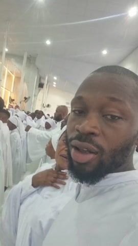 A man takes a selfie in what appears to be a church, judging by the crowd of people in white robes and the building's interior. The man is African, with a beard and short-cropped hair. He is wearing a white robe as well. He makes a few faces for the camera: sticking out his tongue, opening his mouth wide, and pursing his lips. There is a woman sitting behind him, also in a white robe, who smiles at the camera. The building is brightly lit, with tall white columns and multiple lights on the ceiling. There are ceiling fans visible as well. The mood seems reverent and spiritual.