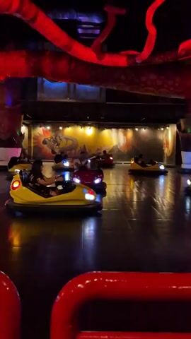 People are riding bumper cars at an indoor amusement park. The cars are mostly yellow and red. Red lighting illuminates the ceiling. The floor is reflective black. There are murals in the background. A red metal barrier surrounds the ride.