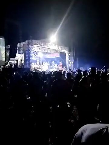 A live outdoor concert is taking place at night. A large crowd of people are gathered in front of a stage. The stage is illuminated by bright lights, and there are several musicians performing. A large screen is set up to the left of the stage, displaying a video feed of the performance. The overall atmosphere appears to be energetic and lively, with the audience actively engaged in the event.