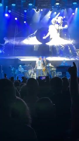 The video shows a live concert performance by the band Deep Purple. In the foreground, a crowd of concert-goers is visible, with many holding up their phones to record. In the background, the band members are on stage performing. Two lead singers are in the center, one in a white t-shirt and the other in a dark shirt and jeans playing a guitar. Behind them, a drummer with the words "DEEP PURPLE" on his drum kit, a keyboard player, and another musician are visible. The stage is lit with bright, colorful lights, and a large screen behind the band displays abstract visuals, including what appears to be a spinning vinyl record and a mirrored kaleidoscopic image of playing cards. The overall atmosphere is energetic and celebratory, typical of a rock concert.