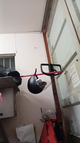 A black motorcycle helmet with a clear visor hangs by its strap from a red bar. The red bar is suspended by a black apparatus that appears to be attached to a larger, red, mechanical device. The helmet and bar are suspended in front of a white wall. To the left of the helmet, a portion of another black object, possibly a motorcycle, is visible. To the right of the helmet, a white vertical surface with some text is attached to the wall. The scene appears to be indoors, possibly a garage or workshop.