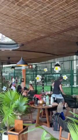 The video depicts a restaurant or bar area next to an indoor padel tennis court. People are seated at tables, some eating and drinking, while others watch a padel tennis game in progress. The restaurant has a thatched roof, hanging light fixtures, and potted plants. The tennis court is enclosed with green walls and netting. Several players are visible, engaged in the game. People are moving around in both spaces.
